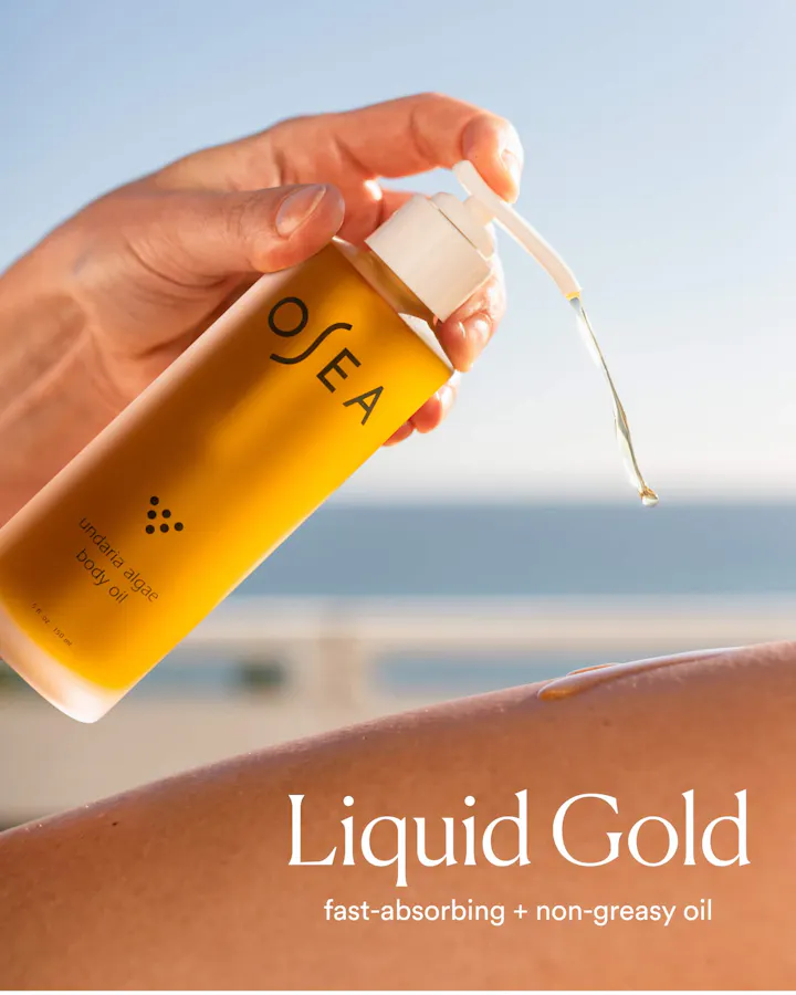 A hand pumping OSEA Undaria Algae Body Oil, with a golden oil droplet falling onto sun-kissed skin against a beach backdrop, with text reading 'Liquid Gold: fast-absorbing + non-greasy oil.