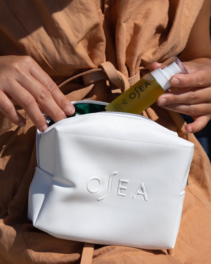 Person placing OSEA skincare bottle into white zippered cosmetic bag for travel