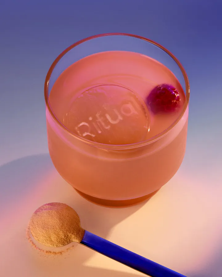 Glass of pink Ritual drink with branded ice cube, cherry garnish, and powder scoop on gradient background