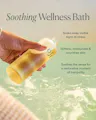 Hand holding a bottle labeled 'Soothing Wellness Bath' with text about its benefits on a blurred bath scene background.