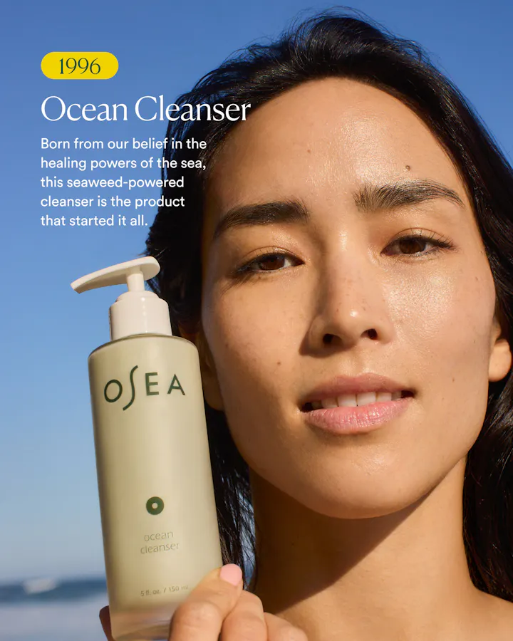 A woman outdoors against a blue sky holds a bottle of OSEA Ocean Cleanser. The bottle is a pale warm beige with a white pump, and the image includes marketing text and a yellow '1996' badge.