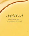 Close-up of a yellow body oil with 'Liquid Gold' text on a beige background