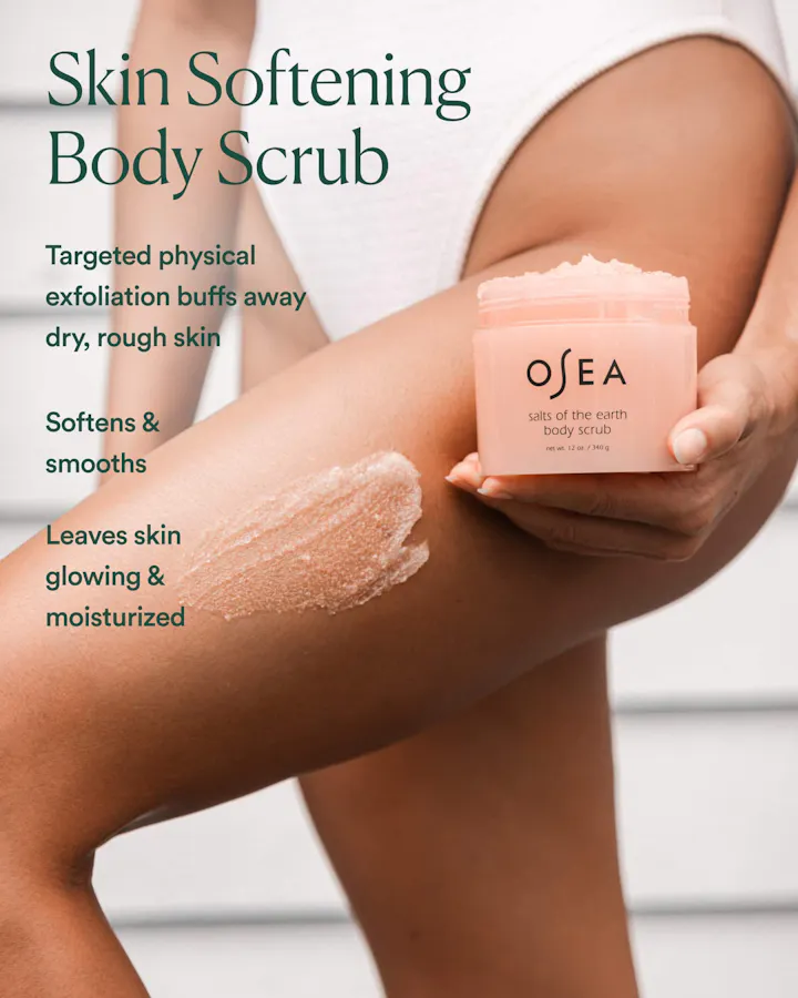 A hand holding an open jar of OSEA Salts of the Earth Body Scrub against a bare thigh with scrub texture swatched on skin, with text reading 'Skin Softening Body Scrub: Targeted physical exfoliation buffs away dry, rough skin. Softens & smooths. Leaves skin glowing & moisturized.'