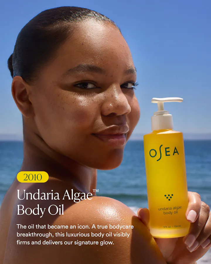 A woman outdoors by the ocean holds a yellow OSEA Undaria Algae Body Oil bottle. The image includes promotional text on the left side.