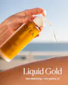 A hand holds a frosted bottle of OSEA undeniably body oil, with golden oil being dispensed, against a blurred beach background. Text at the bottom reads 'Liquid Gold fast-absorbing + non-greasy oil'.