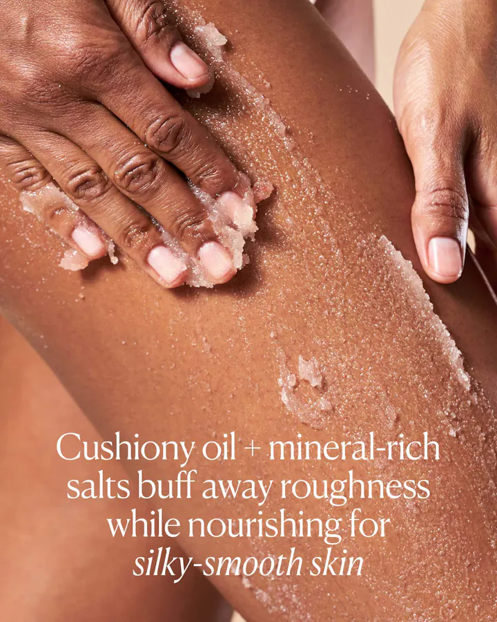 Close-up of hands massaging OSEA Salts of the Earth Body Scrub into skin, showing the gritty salt-and-oil texture, with text reading 'Cushiony oil + mineral-rich salts buff away roughness while nourishing for silky-smooth skin.