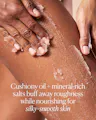 Close-up of hands massaging OSEA Salts of the Earth Body Scrub into skin, showing the gritty salt-and-oil texture, with text reading 'Cushiony oil + mineral-rich salts buff away roughness while nourishing for silky-smooth skin.