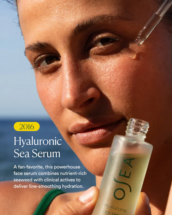 Close-up of a woman applying hyaluronic sea serum with a glass dropper, with the OSEA bottle held in her hand against a blue sky background.