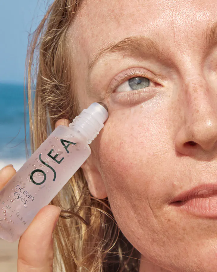 Person applying a skincare product labeled 'Ojea' to their eye with a beach in the background