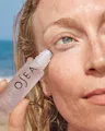 Person applying a skincare product labeled 'Ojea' to their eye with a beach in the background