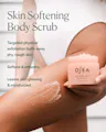 A person holding an open jar of OSEA Skin Softening Body Scrub while applying a beige scrub to their leg. The image features dark green text detailing the benefits of the body scrub on the left side.