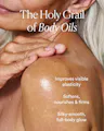Close-up of a hand smoothing oil onto a shoulder, with text reading 'The Holy Grail of Body Oils: Improves visible elasticity, softens, nourishes & firms, silky-smooth full-body glow.'