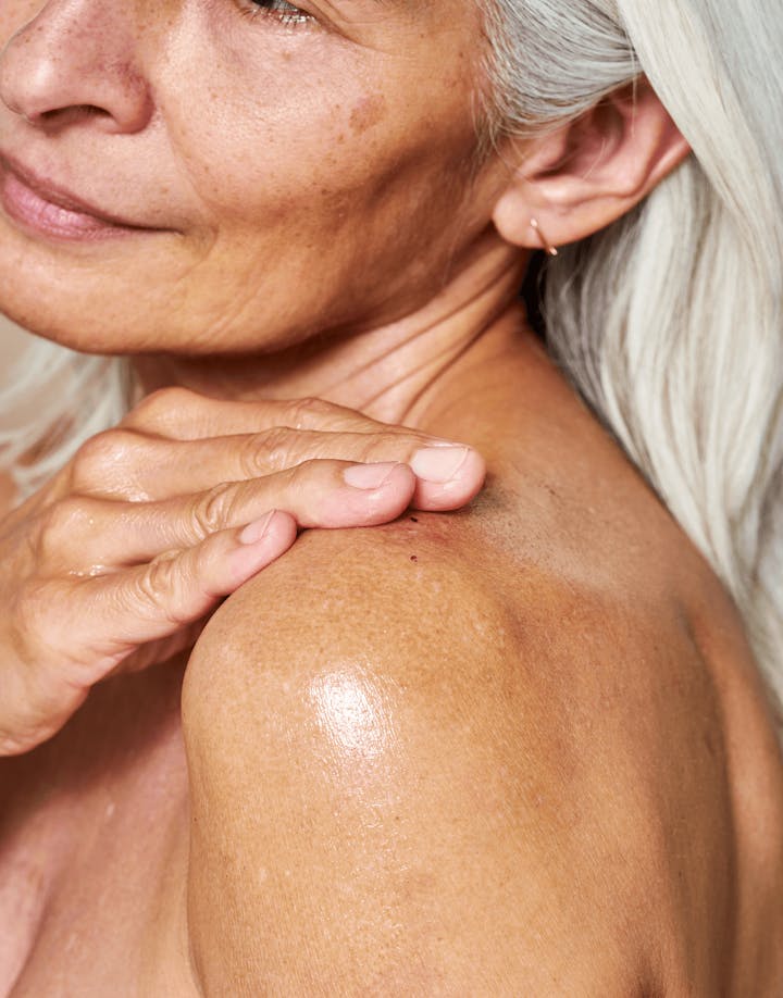 Mature model applying body moisturizer to shoulder, demonstrating visible hydration and skin softness