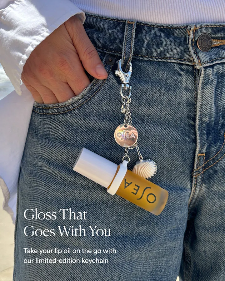 A close-up lifestyle shot of a person wearing blue jeans and a white top, showing a silver O'nea keychain clipped to the waistband. The keychain includes a small gold O'nea lip oil bottle charm, a white seashell charm, and a round metallic charm.