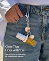 A close-up lifestyle shot of a person wearing blue jeans and a white top, showing a silver O'nea keychain clipped to the waistband. The keychain includes a small gold O'nea lip oil bottle charm, a white seashell charm, and a round metallic charm.