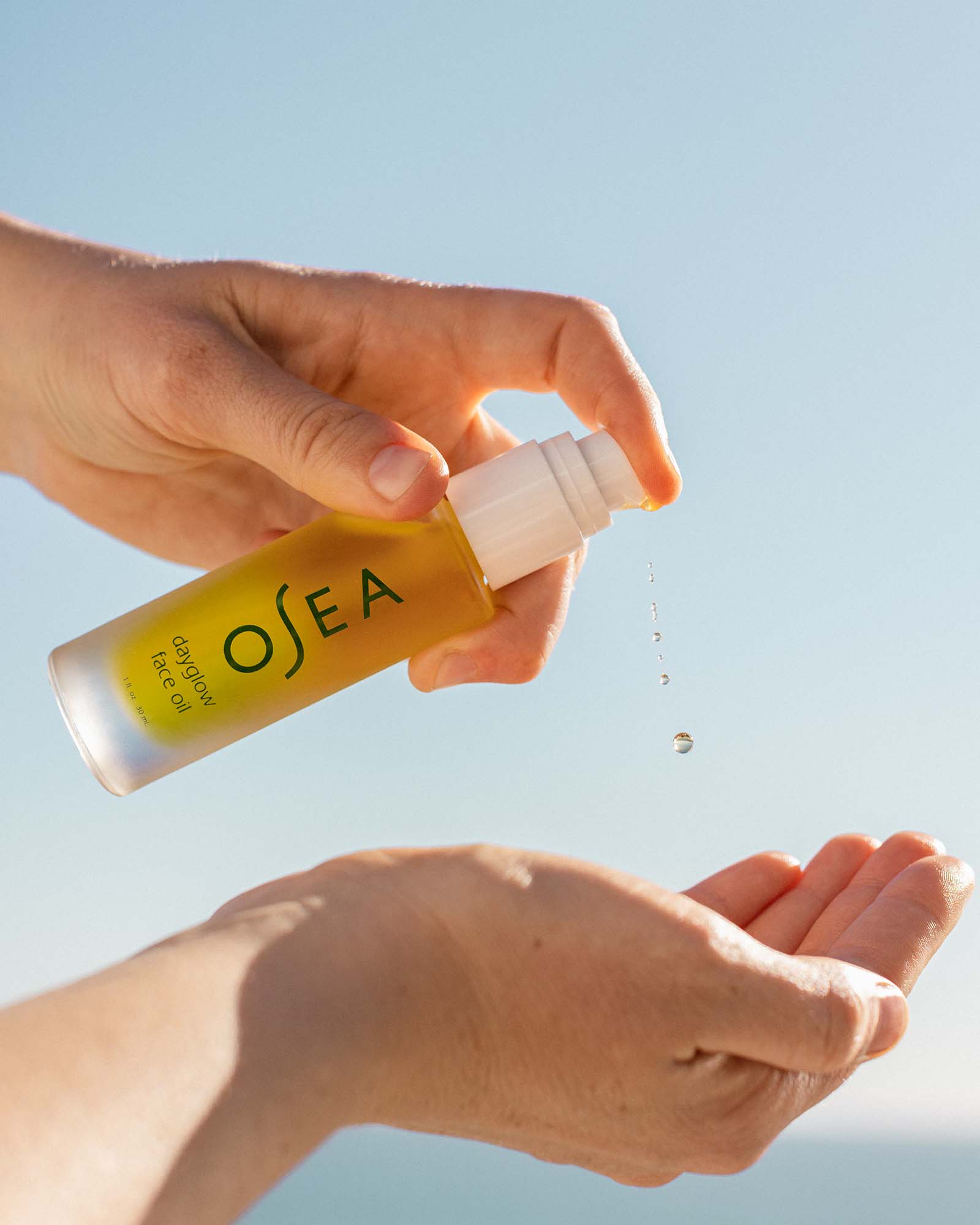 Hand holding a bottle of OSEA Dayglow Face Oil product with a clear blue sky background