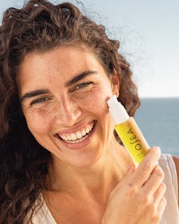 Woman applying a skincare product to her face with a clear blue sky in the background
