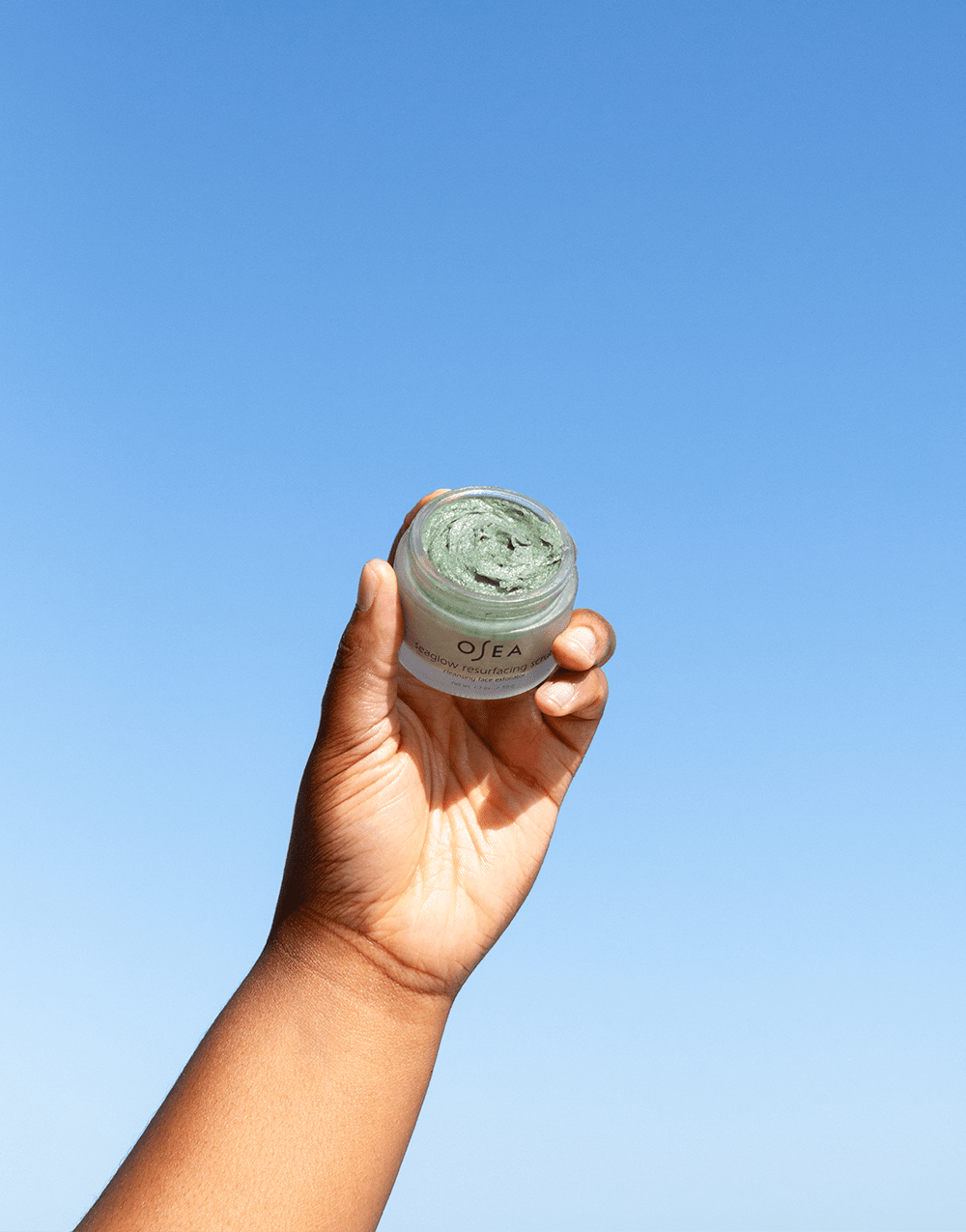 Model raising OSEA Seaglow Scrub jar outdoors with clear blue sky behind