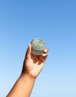 Model raising OSEA Seaglow Scrub jar outdoors with clear blue sky behind