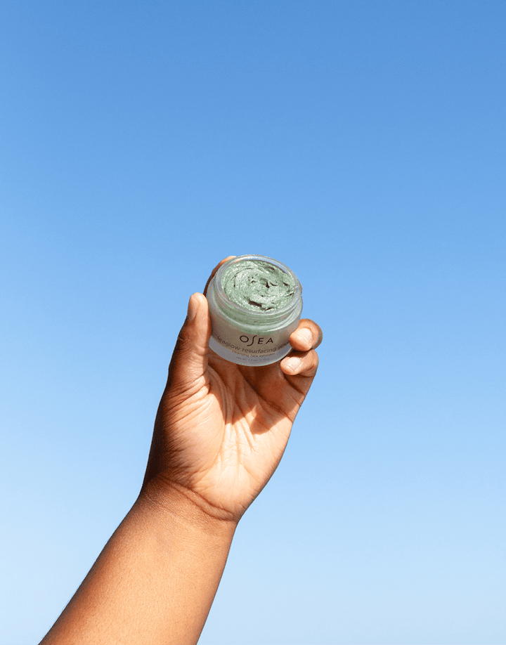 Model raising OSEA Seaglow Scrub jar outdoors with clear blue sky behind