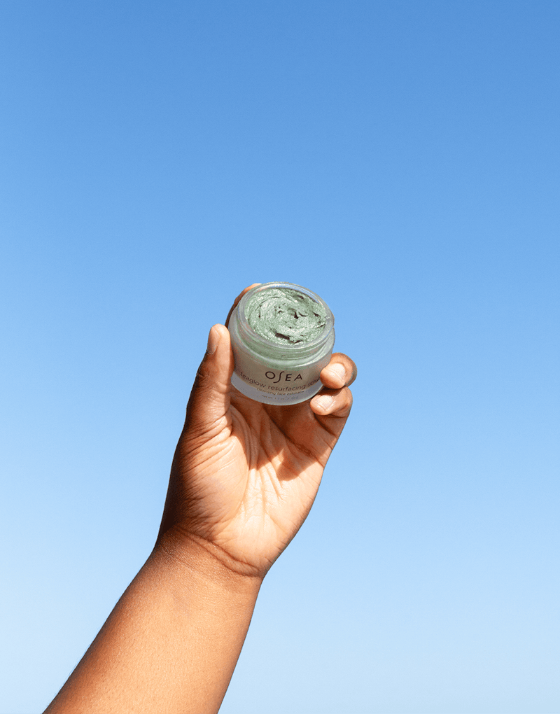 Model raising OSEA Seaglow Scrub jar outdoors with clear blue sky behind