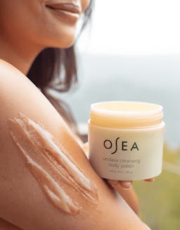 Model applying OSEA Body Butter to shoulder while holding jar, captured in soft natural light