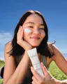 Woman applying sunscreen on her face with a bottle of OSEA product in a grassy outdoor setting.