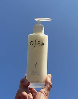 Hand holding OSEA Anti-Aging Body Balm bottle upright against blue sky