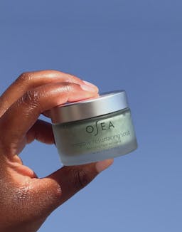 Model holding OSEA Seaglow Resurfacing Scrub jar in natural light against sky background