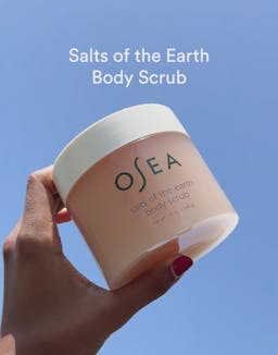 Hand holding OSEA Salts of the Earth Body Scrub jar against blue background