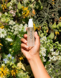Hand holding Essential Hydrating Oil against sunlit garden backdrop