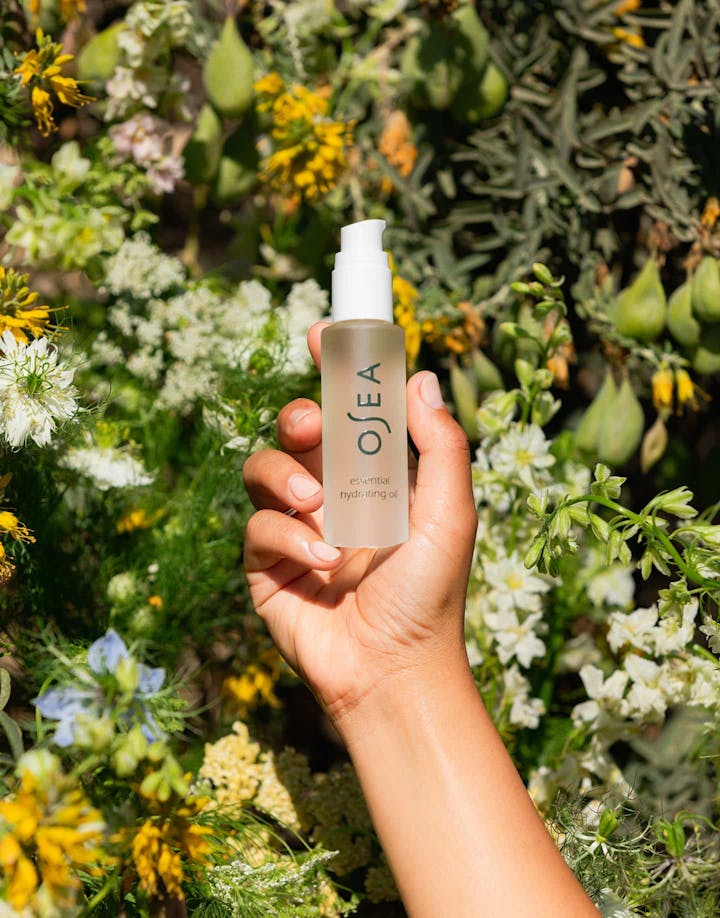 Hand holding Essential Hydrating Oil against sunlit garden backdrop