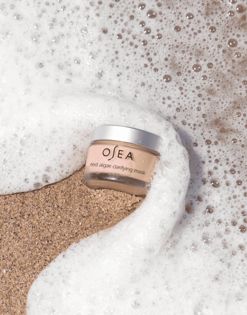 Red Algae Clarifying Mask jar placed in sand with ocean foam surrounding it